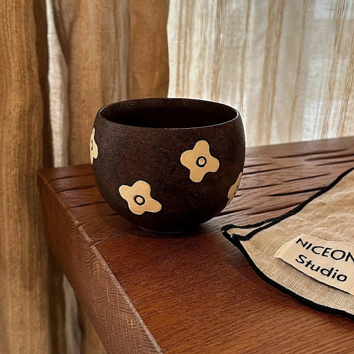 Handmade ceramic mug with gold flower accents, rustic dark glaze, on wooden table near studio pouch