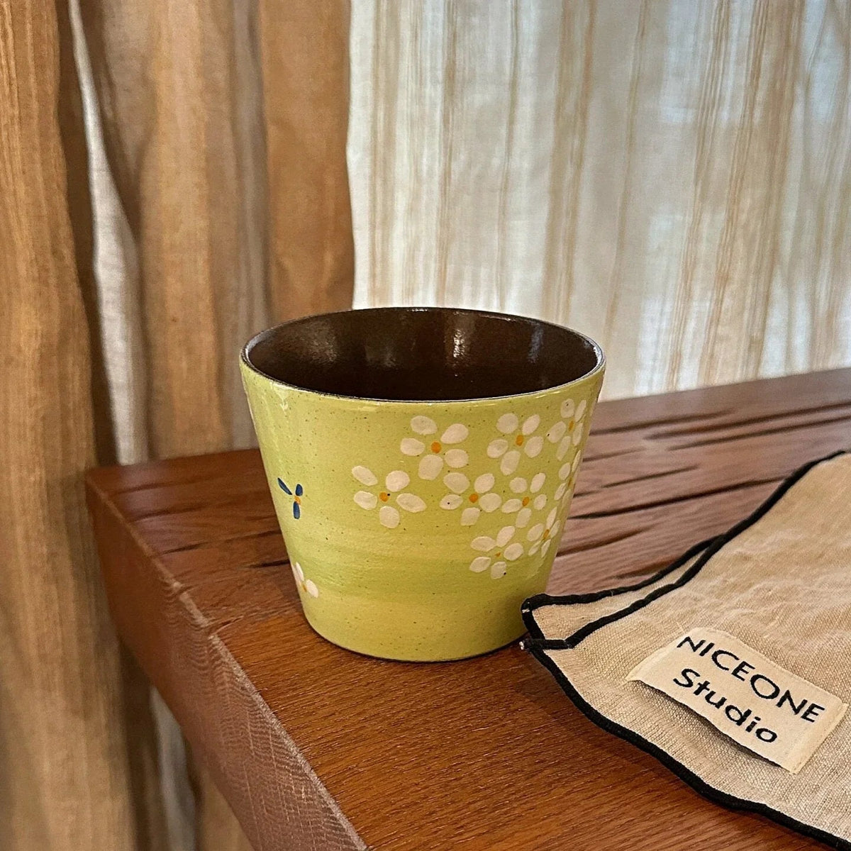 Handmade ceramic mug with white daisy pattern, green glaze, and brown interior, displayed on wooden shelf