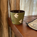 Green ceramic mug with white bird and mushroom designs on wooden table