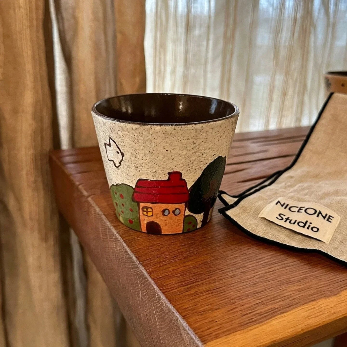 Handmade ceramic mug with cozy house illustration, speckled beige glaze, and brown interior, displayed on wooden shelf.