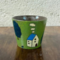 Handmade ceramic mug with green glaze, blue-roofed house, tree, and clouds design, perfect for coffee or tea.