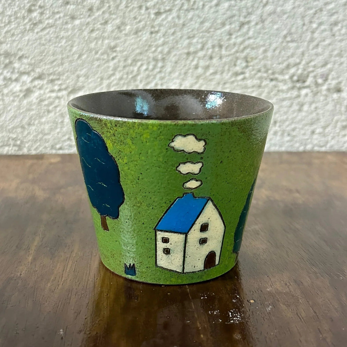 Handmade ceramic mug with green glaze, blue-roofed house, tree, and clouds design, perfect for coffee or tea.