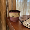 Handmade ceramic mug with green floral pattern, purple rim, and speckled beige band, on wooden table near curtain