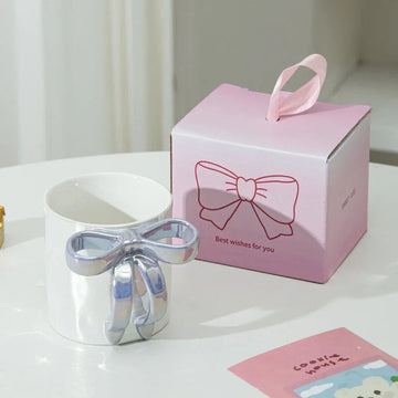 White ceramic mug with shiny silver bow handle beside pink gift box, perfect for tea or coffee