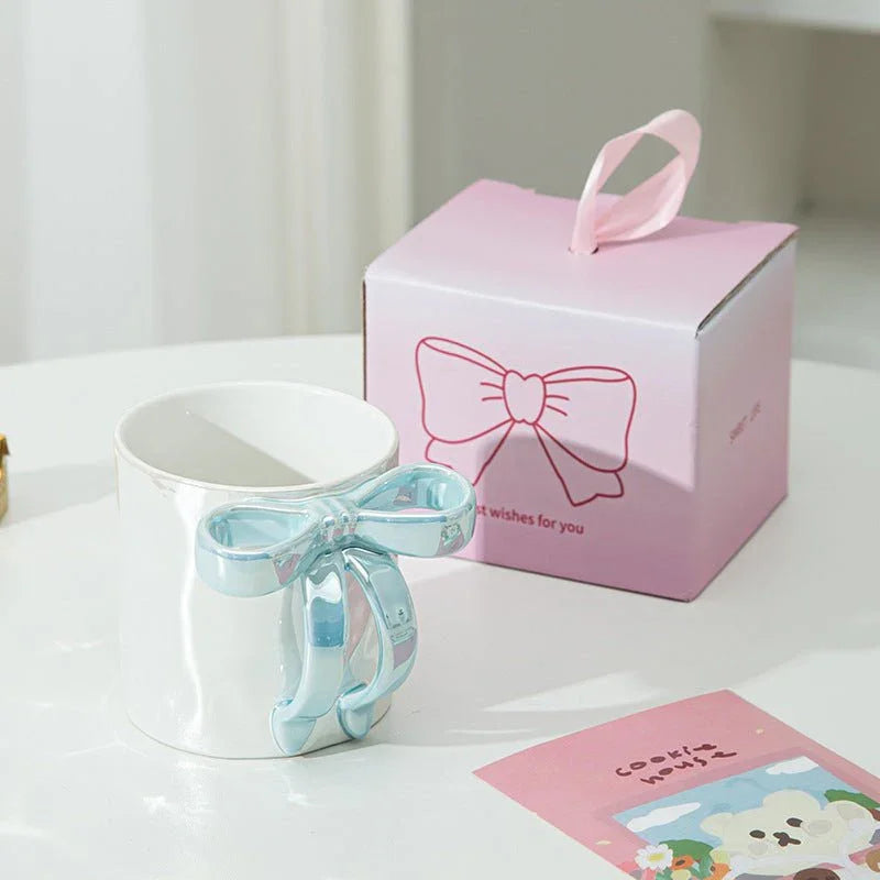 White ceramic mug with blue bow handle beside pink gift box, perfect for tea or coffee lovers