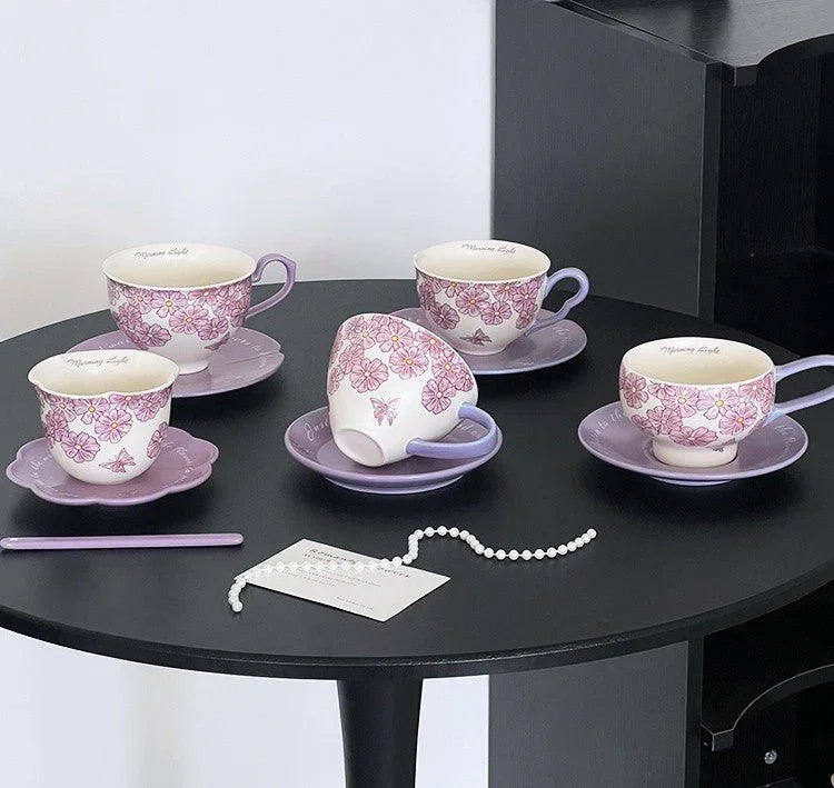 Handmade ceramic tea cups with pink floral design, matching saucers, and pearl necklace on black table