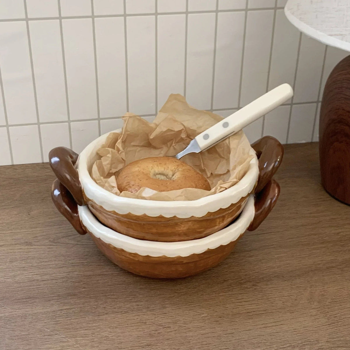 Handmade ceramic bowl with handles, holding a bagel on parchment paper, perfect for breakfast or tableware